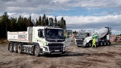 The Volvo FM and Volvo FMX electric trucks off road on muddy ground with trees in the background.