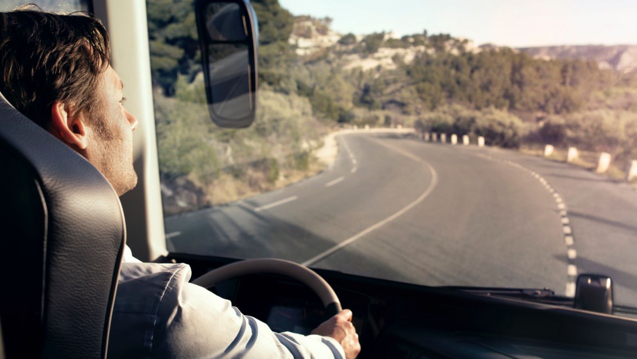 Imagen de un hombre tomada desde detrás de su hombro derecho. Está conduciendo el autobús en una carretera rodeada de colinas con aspecto mediterráneo.