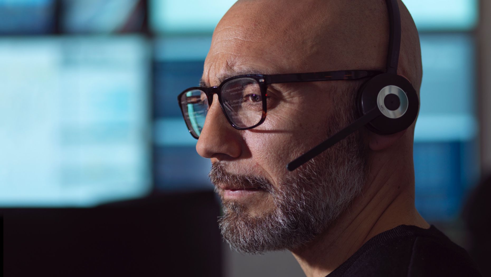 Close-up van een man met een bril en een headset met microfoon die naar een computerscherm kijkt.