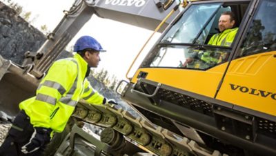 El conductor de una excavadora amarilla de Volvo Group en un sitio de construcción conversando con un compañero de trabajo de pie en el suelo