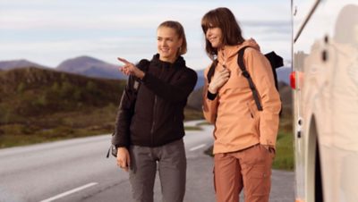 Två kvinnor som pratar bredvid en buss som står parkerad bredvid en bergsväg.