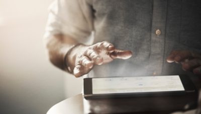 Tronco e braços de uma pessoa atrás de uma mesa com um tablet em cima. As mãos estão próximas à tela.