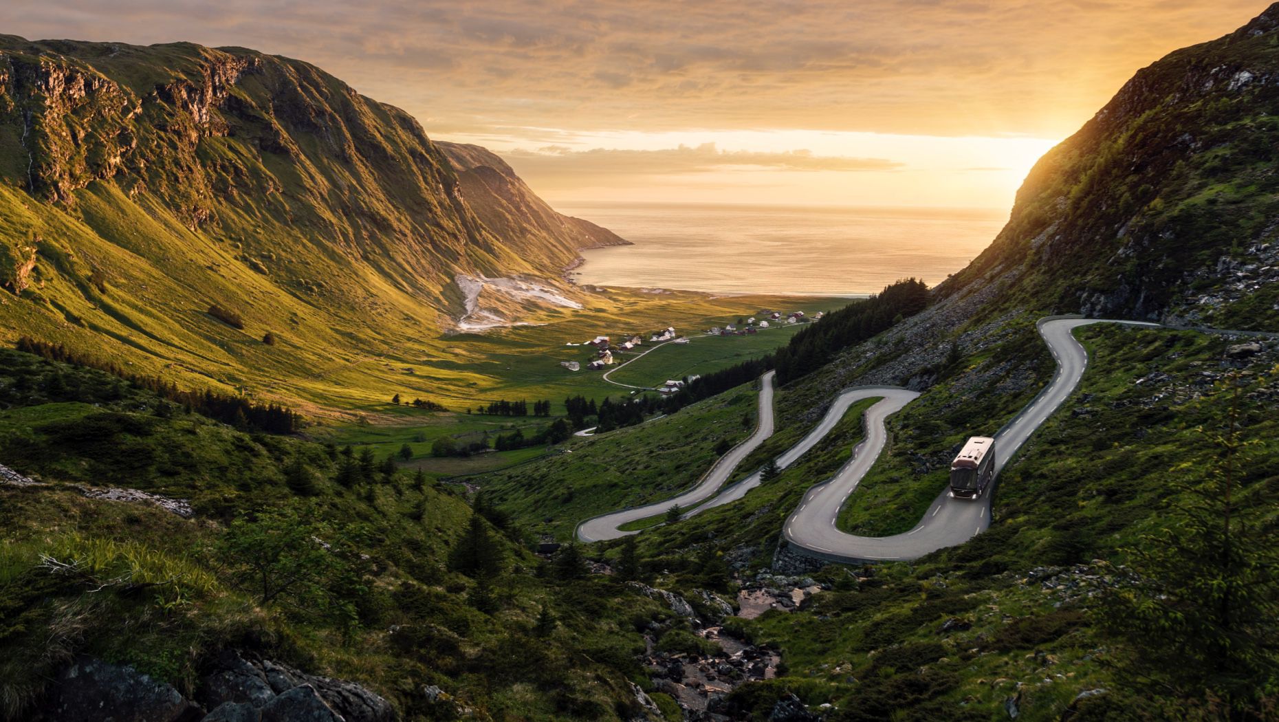 Bus on a meandering road leading down a steep valley towards the sea at dawn.