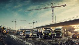 Six Volvo trucks lined up at a construction site beneath a bridge next to two wheel loaders