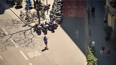 A skateboarder in the middle between two roads with mopeds parked behind him on the street and bikes on the pavement