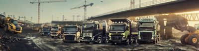 Six Volvo trucks lined up at a construction site beneath a bridge
