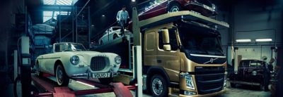 Voiture particulière Volvo Blanche rétro sur un pont élévateur à côté d'un camion Volvo de couleur Bronze transportant deux voitures, une rouge et une blanche