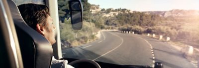 Imagen de un hombre tomada desde detrás de su hombro derecho. Está conduciendo el autobús por una carretera rodeada de colinas de aspecto mediterráneo.