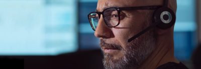 Primer plano de un hombre con gafas y unos auriculares con micrófono mirando el monitor de un ordenador.