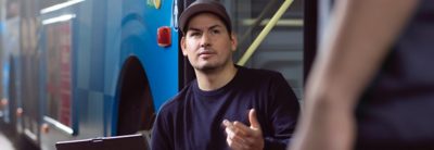 A workshop technician in conversation with unseen person, in front of a bus. 