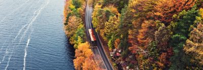 Dubbeldekkerbus op een weg bij een meer en een bos met herfstkleurige bladeren