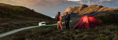 Two hikers in front of their tent, behind a bus on a mountain road