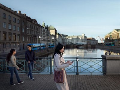 Mulher a atravessar uma ponte sobre um canal numa cidade. Dois peões na periferia. Gráficos sobrepostos na fotografia a mostrar exemplos do sistema de gestão de frota.