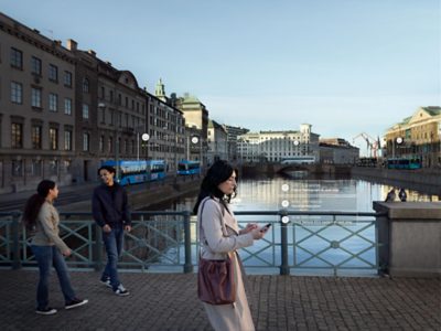 A woman uses Volvo Buses bus fleet management system while crossing a bridge over a city canal. Two pedestrians in the periphery. 