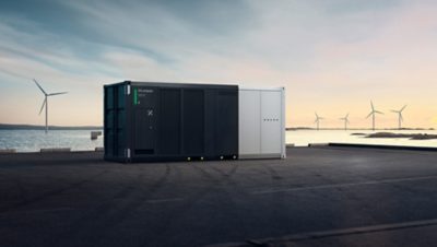 BESS from Volvo Energy stands at a port with the sea and wind turbines in the background.