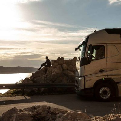 Járművezető egy sziklán ül, kilátással a tengerre naplementekor, mellette parkoló Volvo FH teherautójával