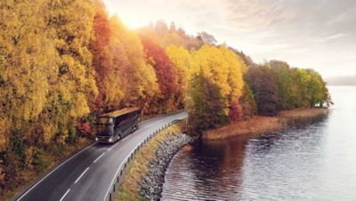 Dubbeldekkerbus op een weg bij een meer en een bos met herfstkleurige bladeren