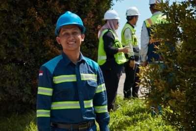 A smiling construction worker wearing a blue hard hat and reflective-striped uniform stands in the foreground.