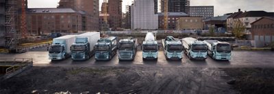 Sähkökäyttöisiä Volvo-tasakuorma-autoja rinnakkain kaupunkimaiseman edessä.