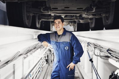 Mechanik sebevědomě stojí v údržbářské jámě pod velkým vozidlem v modrém montérku s viditelným logem Volvo. Prostředím je jasně osvětlená, moderní automobilová dílna s čistými bílými stěnami a viditelným potrubím. Osoba je mužského pohlaví, dospělý a zdá se, že provádí kontrolu nebo opravu vozidla. Celková atmosféra je profesionální a technická, se zaměřením na servis automobilů.