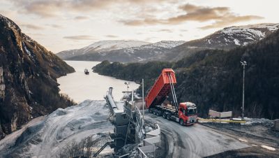 An orange Volvo Autonomous Truck unloading its contents with the backdrop of a water body with a boat on it.