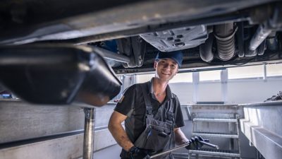 Lächelnde Kfz-Mechatronikerin bei Volvo Trucks steht mit verschränkten Armen selbstbewusst in einer hellen, modernen Werkstatt, im Hintergrund ist ein angehobener Lkw zu sehen