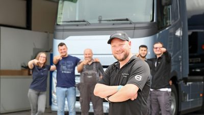 Lächelnde Kfz-Mechatronikerin bei Volvo Trucks steht mit verschränkten Armen selbstbewusst in einer hellen, modernen Werkstatt, im Hintergrund ist ein angehobener Lkw zu sehen