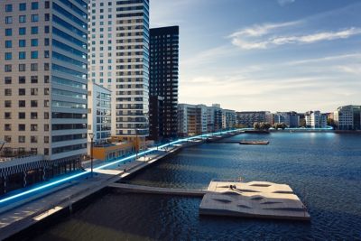 A modern city scene with a blue glowing line visualizing the path of an electric bus along a quay.