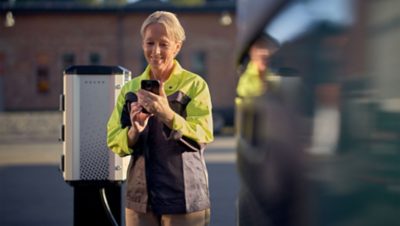 A woman looking at her phone, standing in front of a Volvo Energy charging station.