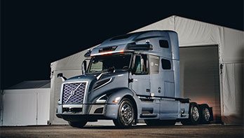Autonomous Volvo semi-truck with the Aurora driver standing in front of a warehouse.