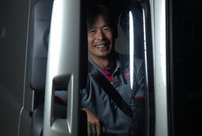 A smiling man sits in the driver’s seat of a vehicle at night, photographed through the side window. 