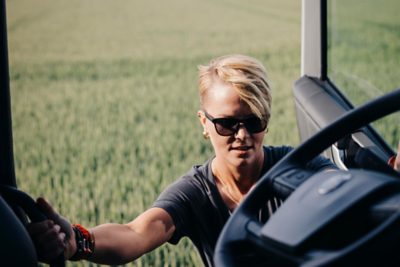 Osoba o krótkich blond włosach i w okularach przeciwsłonecznych wchodzi do kabiny samochodu ciężarowego Volvo, trzymając się wewnętrznych uchwytów, na tle rozmazanego zielonego pola uprawnego.