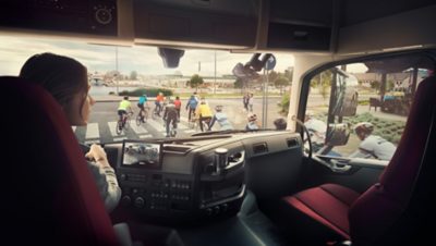 Lkw-Fahrer wartet, während Radfahrer einen Fußgängerüberweg überqueren, sichtbar durch die Windschutzscheibe, Spiegel und Rückfahrkamera.