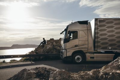 A truck parked behind a driver who is sitting on rocks and looking out over the water