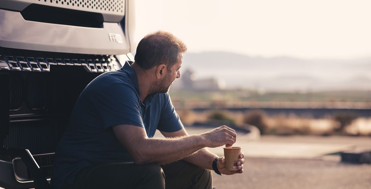 Mannelijke chauffeur zit en rust voor een Volvo FH16 Aero, terwijl hij van een kop koffie geniet