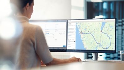 A person sits at a desk in front of a computer screen showing a truck's route on a map