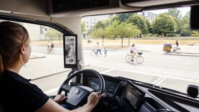 Nő egy teherautó volánjánál egy nagyvárosban, miközben kerékpárok haladnak el mellette