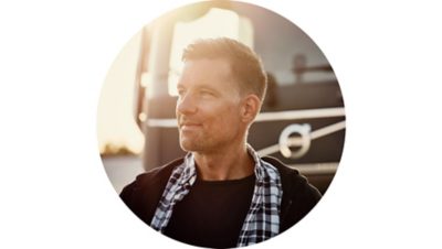 A driver stands in front of a Volvo truck as the sun sets behind him