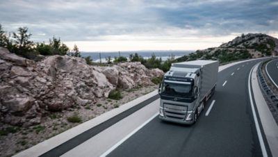 En Volvo FH kjører på en svingete motorvei med steiner og vann i bakgrunnen