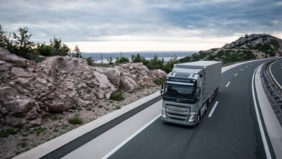 A Volvo FH drives on a curving highway road with rocks and water in the background