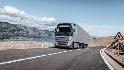 A Volvo FH drives on a highway through a rocky landscape with mountains and water in the background