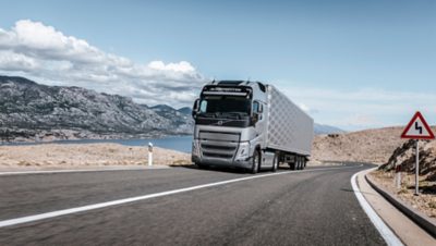 A Volvo FH drives on a highway through a rocky landscape with mountains and water in the background