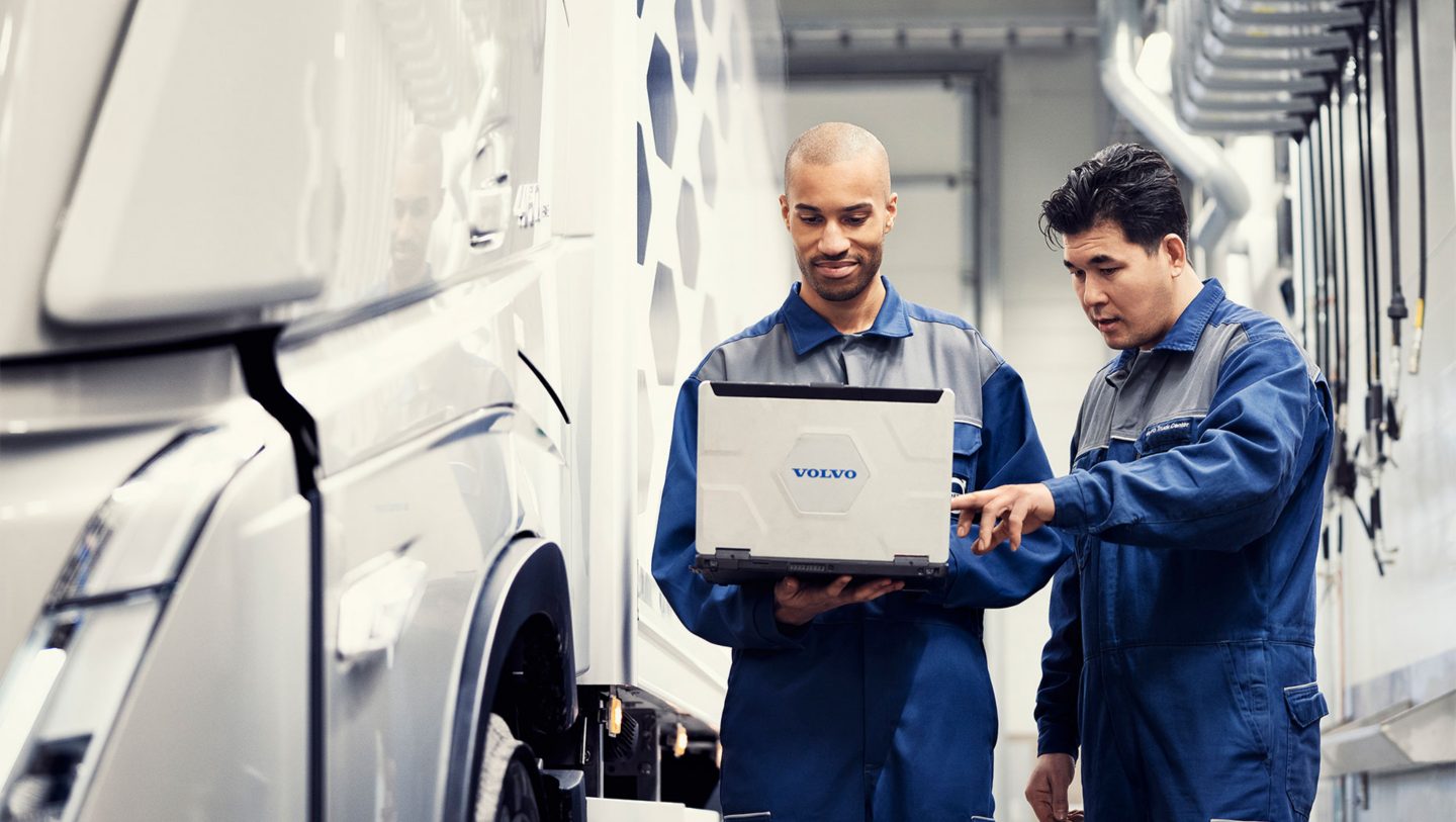 Two Volvo technicians stand next to a truck in the workshop, looking at a laptop