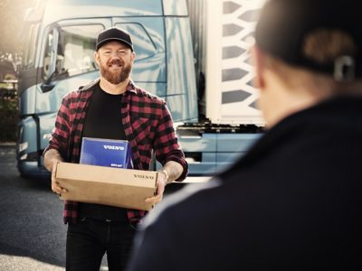 A driver holds a blue Genuine Volvo Parts box as he talks to someone