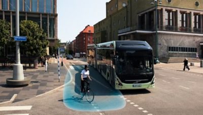 Un autobús urbano eléctrico de Volvo con un ciclista en un campo azul gráfico junto al autobús, para mostrar cómo el sistema de detección lateral del autobús mejora la seguridad vial.