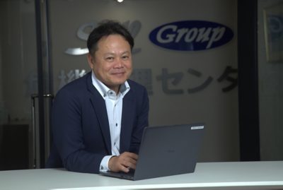 Kazunori Ishitani in a navy blazer and white shirt is seated at a white table, typing on a dark Dell laptop.