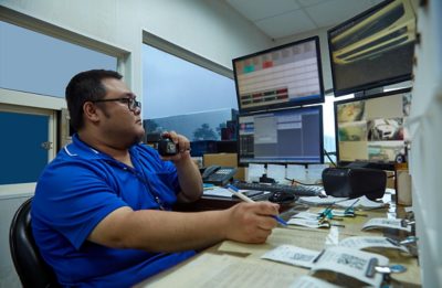 A man in a blue polo shirt sits at a control desk in a monitoring room.