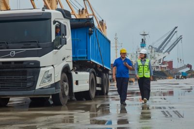 Two workers in hard hats and safety gear walk along a wet dock beside a white Volvo FM truck with a blue trailer.