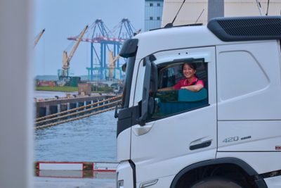 A smiling truck driver looks out the window of a white cab parked near a waterfront.
