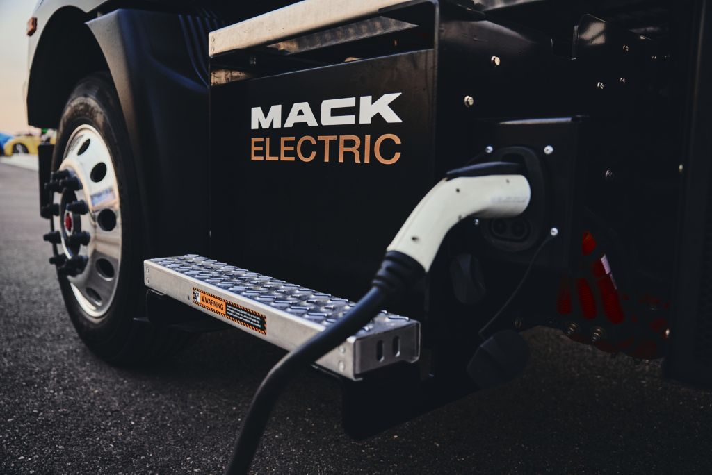 Image shows a charger plugged into an electric Mack . The image only shows the left front wheel and below. On the truck, the words Mack Electric is written one word below the other, towards the right of which the charging port into which the charger is connected.